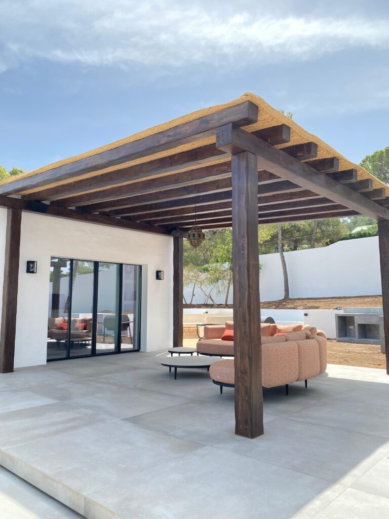 Pérgola de madera oscura adosada a vivienda en Ibiza con mobiliario exterior